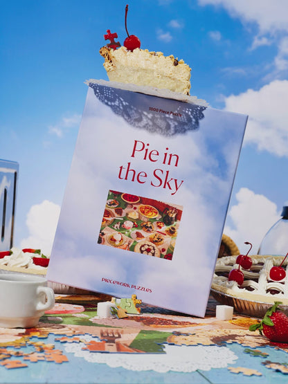 Video showing the front of Piecework Puzzles' Pie in the Sky 1000 piece jigsaw puzzle box. It's a vibrant table scene surrounded by pie and diner coffee. A toy plane and ceramic dog move across the frame during the video.