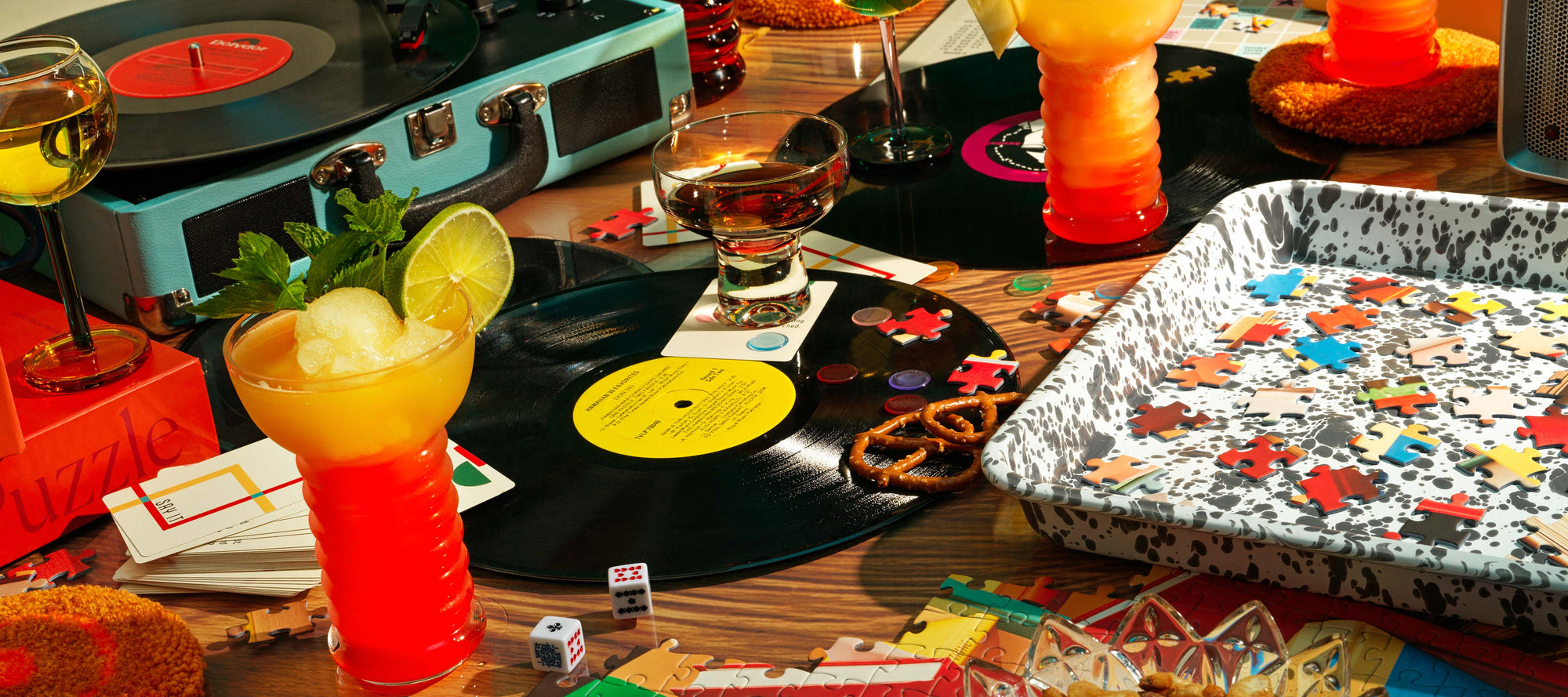 A warm tabletop scene featuring a retro record player, handcrafted cocktails, and a tray filled with colorful puzzle pieces—celebrating the Piecework Playlists experience, where (almost) every puzzle comes with a perfectly paired soundtrack.
