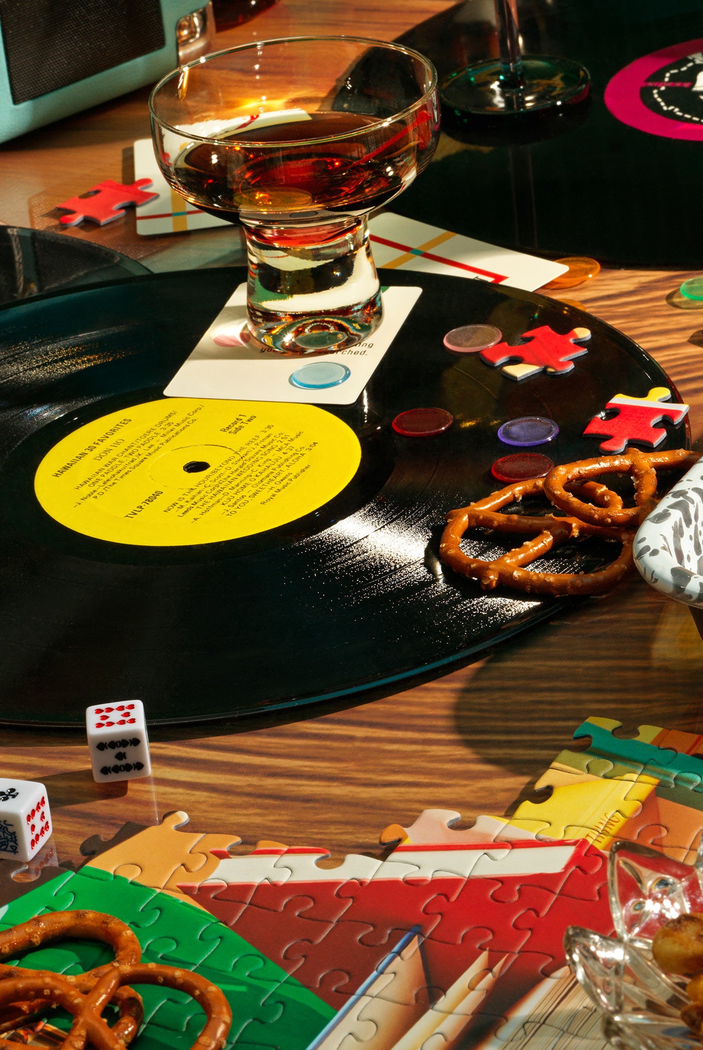 A warm, retro tabletop scene featuring a vinyl record, handcrafted cocktails, and a tabletop filled with colorful puzzle pieces—celebrating the Piecework Playlists experience, where (almost) every puzzle comes with a perfectly paired soundtrack.